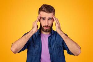 Millennial guy suffering from headache, massaging temples to get rid of pain on orange studio background. Young man having hypertension, feeling stressed or exhausted, cannot find problem solution photo