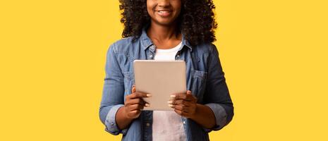 Happy beautiful black woman using modern digital tablet over yellow studio background, copy space. Cute african american young lady testing new mobile application on pad and smiling photo