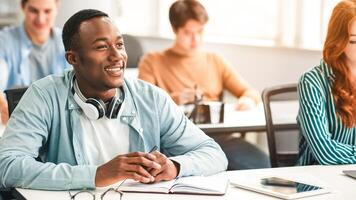 Return To Education Concept, First Day Of Highschool. Portrait Of Smiling African American Male Student Listening To Lecturer at University, Sitting At Desk In Classroom With Multiethnic Classmates photo