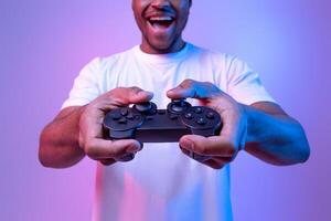 Excited african american guy playing games with joystick, joyful black man enjoying modern entertainment, having fun in vivid neon light on purple studio background, selective focus, copy space photo