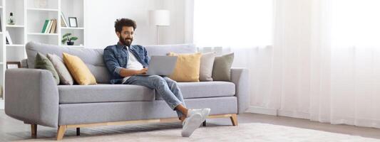 Remote Work. Smiling Indian Freelancer Guy Working With Laptop At Home, Millennial Eastern Man Sitting On Couch In Living Room And Using Computer, Enjoying Distance Job, Panorama With Copy Space photo