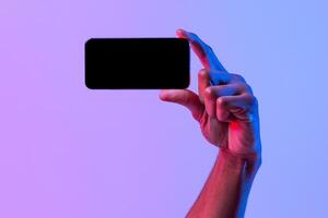Black Male Hand Holding Blank Smartphone In Horizontal Orientation Over Purple Neon Background, African American Man Demonstrating Copy Space For App Or Mobile Game Design, Mockup Image photo