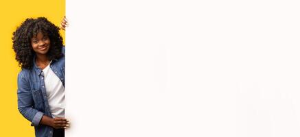 Smiling curly black woman standing by white empty board for advertisement over yellow background. Cheerful african lady with big blank advertisement placard, empty space for text, panorama photo