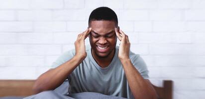 African American Man Suffering From Headache Pain Massaging Temples Having Migraine Sitting In Bed In Bedroom At Home. Healthcare Problem, Painful Head Ache Concept photo
