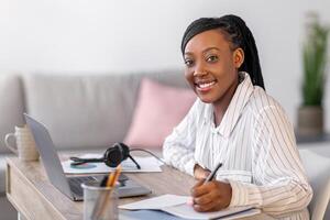 Pretty young black woman enjoying online course, smiling african american lady sitting at workdesk in front of laptop, taking notes in notepad, home interior, side view, copy space photo
