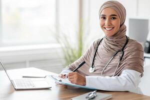 Smiling muslim woman doctor in headscarf taking notes in medical chart, sitting at workdesk with laptop at her cabinet in clinic, taking anamnesis while having conversation with patient, free space photo