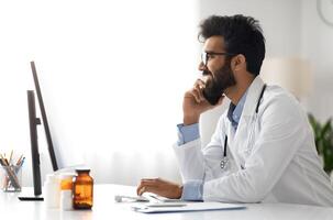 Handsome millennial Arab doctor wearing medical coat working at clinic, general practitioner sitting at workdesk, using computer pc, have phone conversation with patient or colleague, side view photo