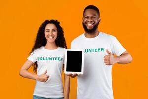 Couple Of Multiracial Volunteers Holding Tablet Computer With Empty Screen Gesturing Thumbs Up Standing On Yellow Background, Smiling To Camera. Studio Shot, Mockup photo