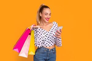 Looking And Searching For Discounts. Smiling female model carrying colorful shopping bags and using smartphone, making purchases in online store standing isolated on orange studio background photo