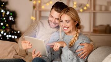 Joyful couple having fun together at home on Christmas, sitting on couch by xmas tree and using digital tablet, copy space. Emotional young man and woman playing game online on pad together photo