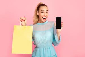 Shopping Application And Online Sales. Overjoyed woman showing mobile phone with black empty screen for mockup to camera and holding bag, standing and posing at pastel pink studio wall background photo