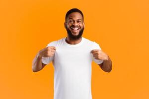 Cheerful Black Man Showing His White T-Shirt Smiling To Camera Standing Over Yellow Studio Background. Look There, Check This Out Concept. T-Shirt Mockup, Template For Your Text. Panorama photo