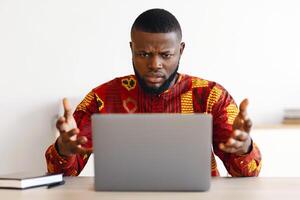 Computer Problems. Frustrated African Businessman In Traditional Clothes Having Troubles With Laptop In Office, Black Guy In Ethnic Shirt Raising Hands Emotionally Reacting To Bad Internet Connection photo