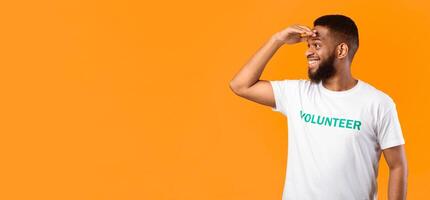African American Volunteer Guy Looking Aside At Copy Space For Advertisement Text Holding Hand Near Eyes Standing Posing Over Yellow Background. Studio Shot photo