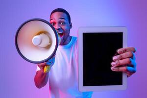 Excited African American Guy With Loudspeaker Holding Digital Tablet With Blank Black Screen, Making Announcement While Standing In Vivid Neon Light Over Purple Studio Background, Mockup Image photo