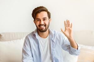 Greeting Concept. Portrait of friendly eastern man waving hello while sitting on couch at home, positive millennial arab guy smiling and looking at camera, saying hello, free space photo