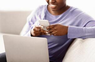 Scrolling News Feed. Closeup of unrecognizable african american smiling lady holding cellphone, typing sms message, sitting on couch with laptop. Black woman browsing internet, surfing web, using app photo