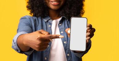 Black woman pointing at smartphone with empty screen in her hand, cropped. Unrecognizable african american lady showing modern mobile phone with blank display, closeup, mockup photo