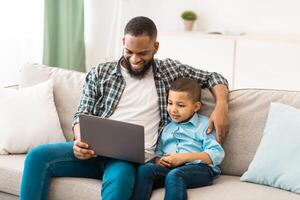 Black Daddy And His Kid Son Watching Movie On Laptop Online Sitting Hugging On Sofa, Spending Weekend Together At Home. Technology And Gadgets, Family Of Two Using Computer In Living Room Indoors photo