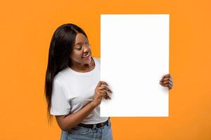 Cheerful african american lady showing empty placard for text or advertisement, orange studio background. Young fair-haired black woman holding blank advertising board and smiling photo
