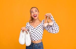 Studio portrait of a smiling stylish woman holding pair of high heels shoes and white sneakers in hands, choosing what to wear, looking up, isolated over pastel orange background wall, copy space photo