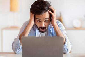 Deadline Stress. Frustrated arab man looking at laptop screen and touching head in shock, eatern freelancer guy suffering business problems while working online on computer at home office, closeup photo
