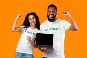 Two Diverse Volunteers Showing Laptop Empty Screen Gesturing Yes Posing Standing Over Yellow Studio Background. Activists Recommending Charity And Volunteering Website. Mockup photo