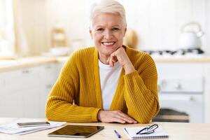 Happy senior woman freelancer working from home, sitting in kitchen, using digital tablet and smartphone, taking notes while reading financial report, smiling at camera, copy space photo