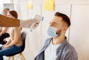 Millennial man in face mask waiting for coronavirus vaccination at health centre, nurse checking his temperature with digital thermometer. Immunization against covid-19 concept photo
