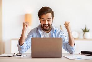 Excited arab guy celebrating success with laptop computer at home office, emotionally reacting to good news, sitting at desk, looking at computer screen, raising fists and exclaiming with excitement photo