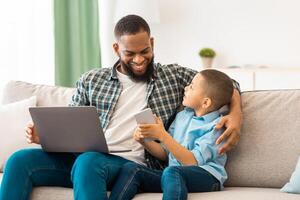 African American Father And Son Using Smartphone And Laptop Sitting On Sofa Indoors. Mobile Application. Family Using Gadgets, Computer And Phone, Browsing Internet At Home. Selective Focus photo