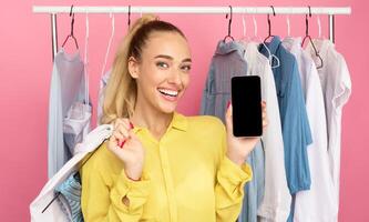 Shopping App Concept. Portrait Of Excited Stylish Woman Showing Cellphone With Black Empty Screen For Mockup And Holding Hanger With Cloth, Standing On Pastel Studio Wall With Rack In Background photo