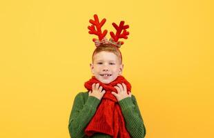 Cheerful kid in green winter sweater and red scarf posing on yellow studio background, copy space. Happy ginger boy with deer accessory on his head waiting for Christmas and New Year photo
