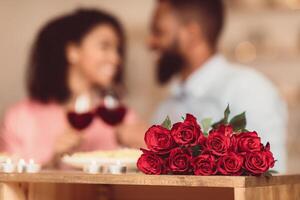 Bonding Concept. Bouquet of flowers lying on table, selective focus on red roses. Happy black man and woman spending time together at home or cafe, having dinner, drinking wine in blurred background photo