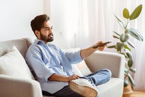 Arab man with remote controller switching channels while watching tv at home, young eastern guy relaxing on couch in comfortable living room, enjoying weekend pastime, copy space photo