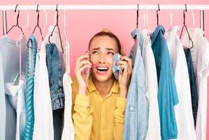 Panic In Wardrobe. Portrait of depressed lady talking on smartphone and crying because she has nothing or can't choose what to wear, wiping tears with shirt, standing in clothes rack between hangers photo