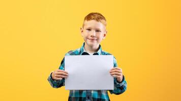Cheerful redhead boy with white blank board on yellow studio background. Cute redhead kid in plaid shirt showing empty placard for your text or advertisement, copy space for advertising photo