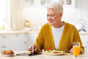 Smiling senior grey-haired woman eating healthy food and using digital tablet in kitchen, empty space. Happy grandmother with glass of fresh juice reading culinary blog on modern pad photo