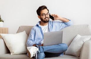 Remote Job Opportunities. Eastern Freelancer Man Talking On Cellphone And Using Laptop At Home, Smiling Arab Millennial Guy Working Online With Computer While Sitting On Couch In Living Room photo