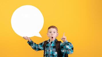 Excited redhead schoolboy with backpack thinking and holding communication bubble, pointing finger up, having great idea, yellow studio background, copy space. Smart kid sharing exciting idea photo