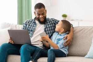 Black Father And Son Using Gadgets, Boy Showing Smartphone To Excited Dad Sitting With Tablet Computer And Browsing Internet Together At Home. Technology, Devices And Modern Family Lifestyle photo