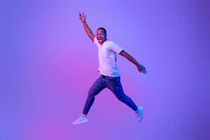 Excited Funny Black Guy Jumping Up In Neon Light Over Purple Background, Full-Length Portrait In Motion Of Euphoric African American Man Jump In The Air With Open Mouth And Raised Hands, Copy Space photo
