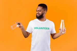 No More Plastic. African Volunteer Guy Choosing Between Glass And Plastic Bottle Standing Over Yellow Studio Background. Ecology And Environment Concept photo
