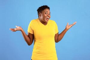 Funny Clueless Oversized Black Lady Shrugging Shoulders Gesturing I Don't Know Posing Standing Over Blue Background In Studio, Looking At Camera. Shoulder Shrug Concept photo