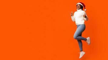 Holiday Offer. Joyful Black Santa Lady In Christmas Hat Jumping And Showing Thumb Up Gesture, Recommending Something, Having Fun On Orange Background In Studio, Cheering Xmas And New Year, Copy Space photo