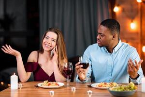 Annoyed African Man Looking At His Girlfriend Talking On Mobile Phone During Romantic Dinner In Restaurant, Complaining About Her Cellphone Addiction. Interracial Couple Having Communication Problems photo