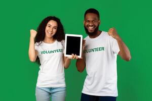 Two Multiethnic Volunteers Showing Digital Tablet Empty Screen Gesturing Yes Celebrating Success Standing Over Green Studio Background, Smiling To Camera. Activists Recommending Volunteering Website photo