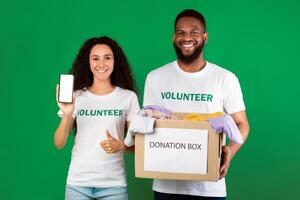 Two Diverse Volunteers Holding Donation Box Showing Smartphone With Blank Screen Picking Clothes For Second-Hand Use Standing Over Green Studio Background, Gesturing Like. Charity And Contribution photo