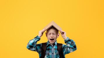 Stressed schoolboy with backpack covering head with book and yelling on yellow studio background, got tired of studying, need for rest, copy space. Ginger boy holding book on his head and screaming photo