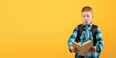 Amazed schooler reading book on yellow studio background. Redhead schoolboy with backpack holding book, having excited face expression, copy space. Books reading, education concept photo